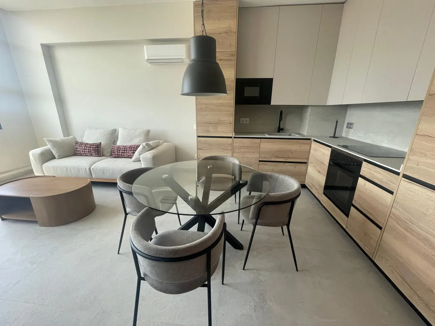 Open-concept apartment with a glass table, gray chairs, a beige sofa, and a modern kitchen with wood-grain cabinets.