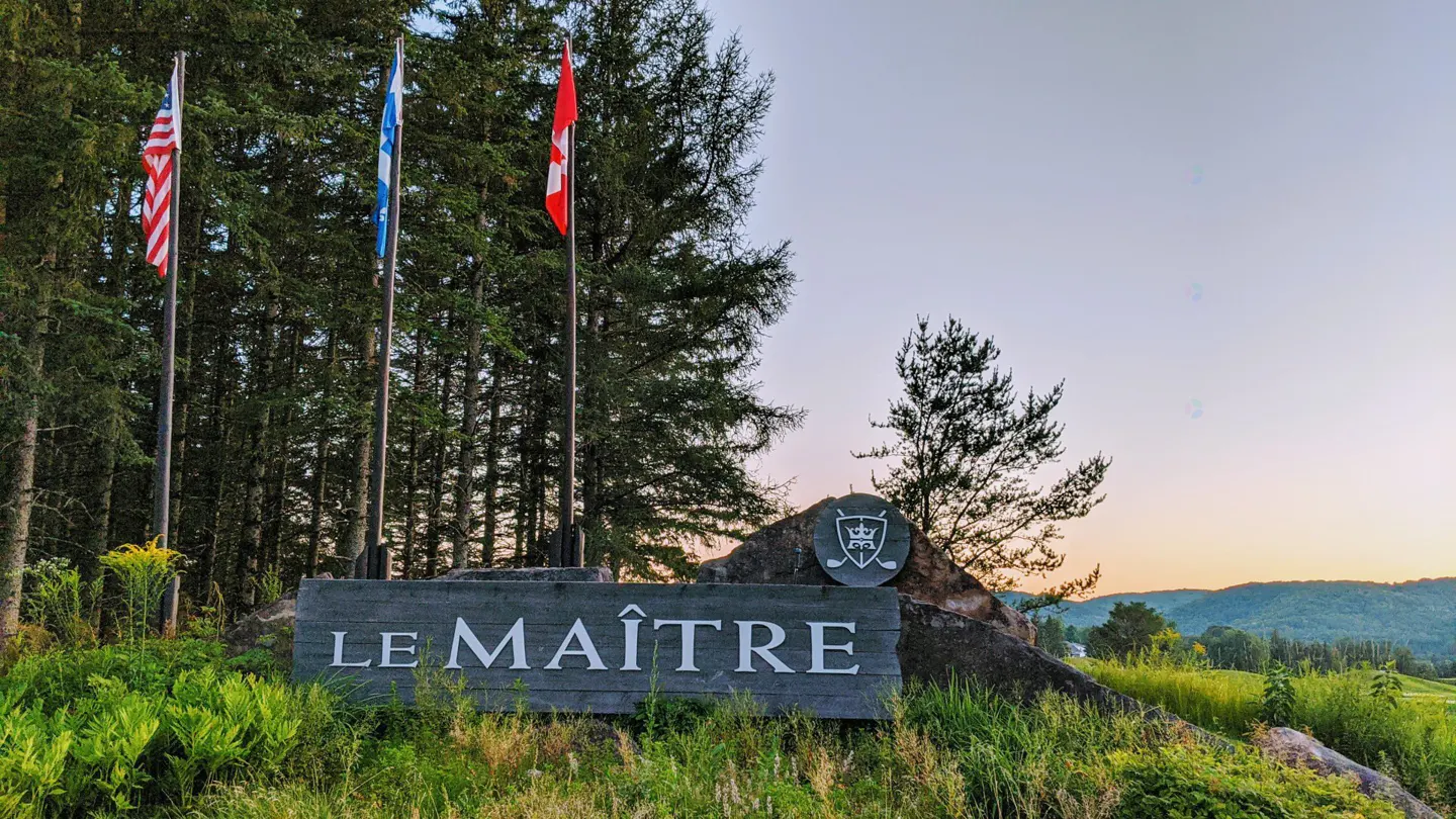 Entrance to Le Maître golf course with US, Quebec, and Canadian flags. Sign is gray wood with white letters. Trees and hills in background.