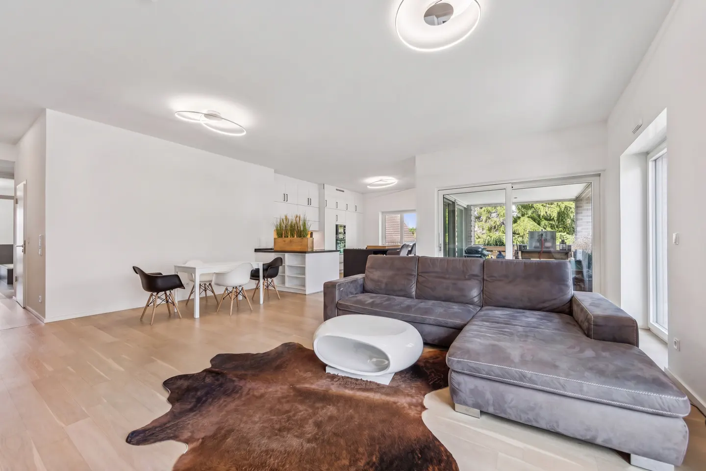 Bright, modern living room with a gray sectional sofa, white table, and cowhide rug on a light wood floor. White walls and circular ceiling lights.