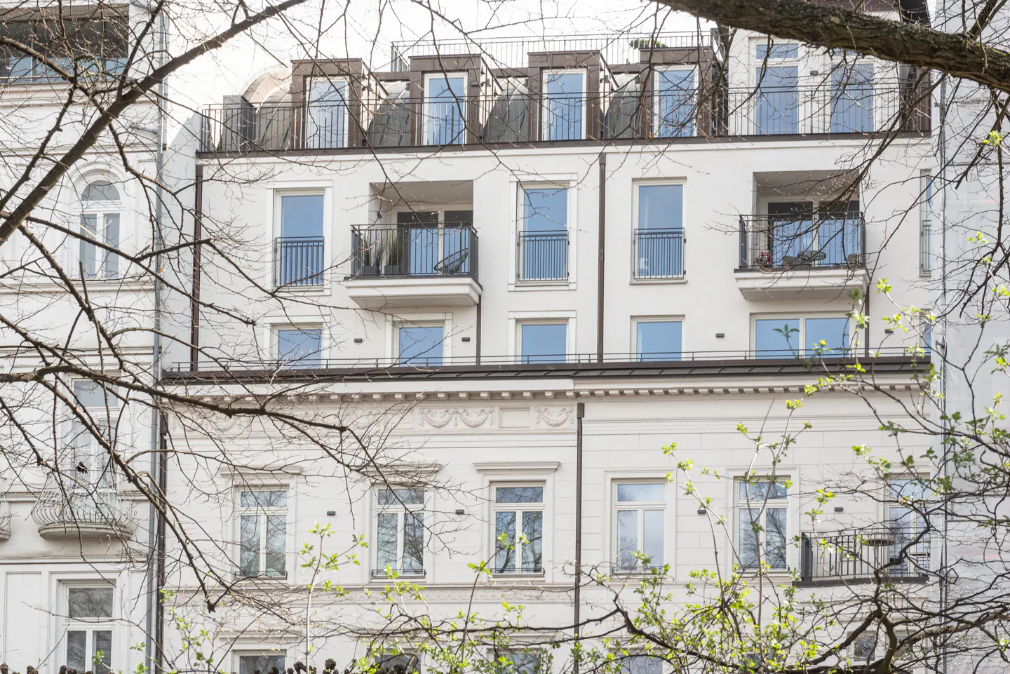 Exterior of a white apartment building with balconies and windows, framed by tree branches.