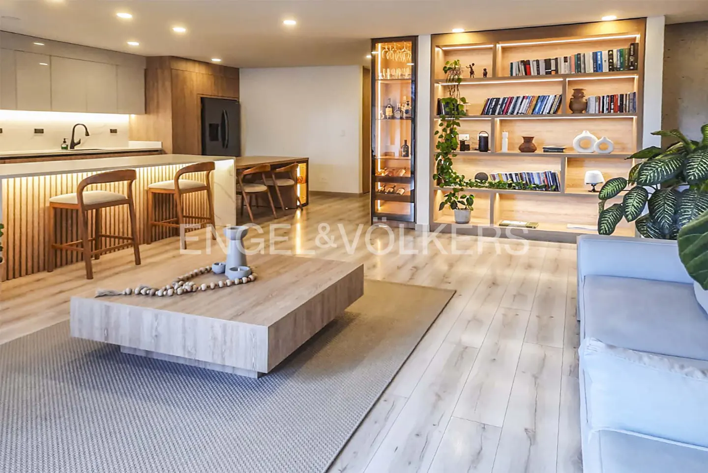 Modern open-plan living area with a wooden coffee table, kitchen island with bar stools, and illuminated bookshelf.