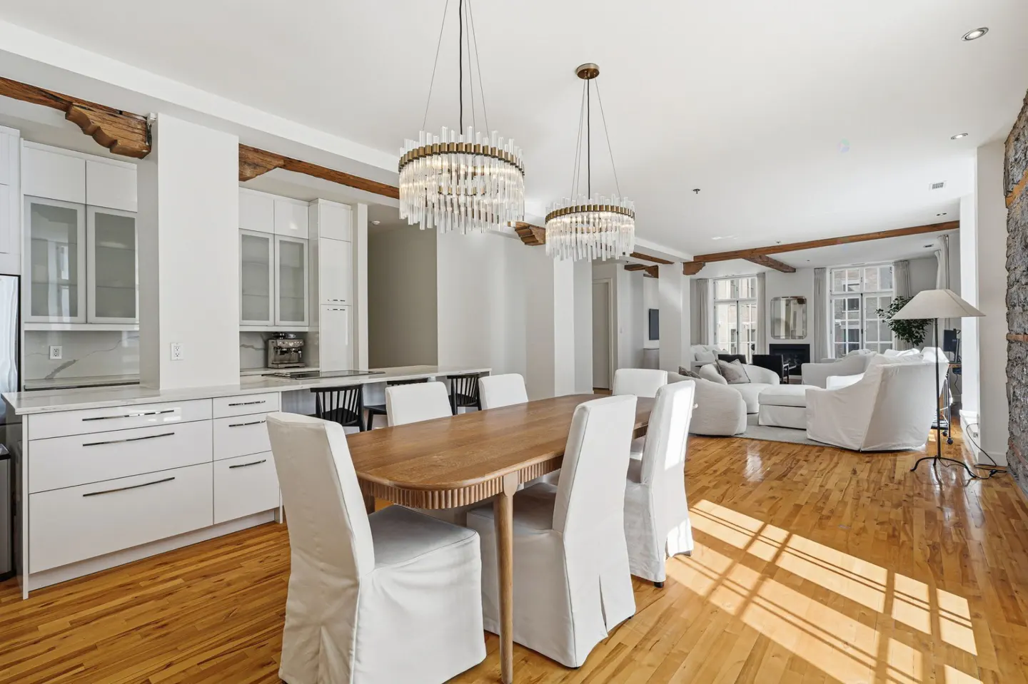Bright, open-concept living space with hardwood floors, white kitchen, and dining table with white-covered chairs. Two crystal chandeliers hang above.
