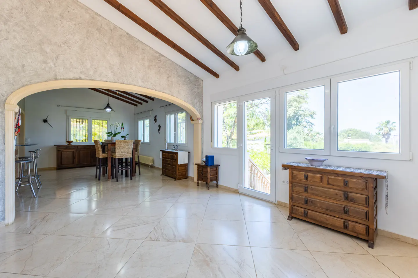 Bright, open-plan living space with marble floors, wooden beams, and an arched doorway leading to a dining area. Large windows offer outdoor views.