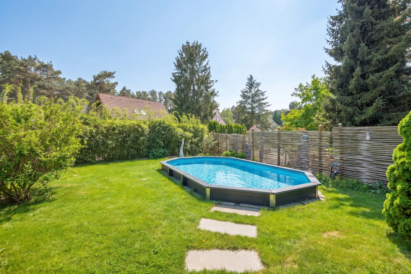Backyard with a blue above-ground pool, stone path, green grass, trees, and a house in the background.
