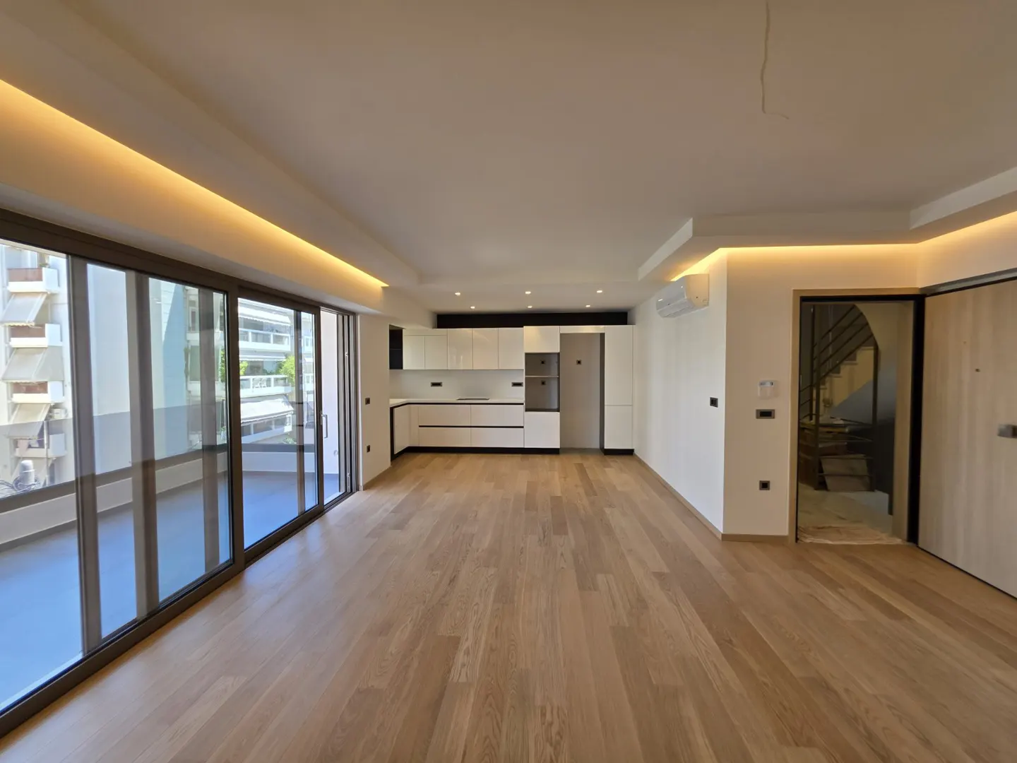 Modern open-plan living area with light wood floors, white kitchen cabinets, recessed lighting, and large glass balcony doors.