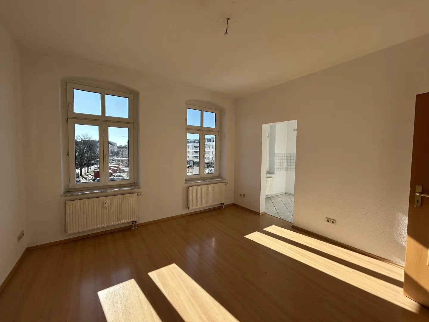 Bright, empty room with wood floors, white walls, and two windows showing a city view. A doorway leads to a white-tiled kitchen.