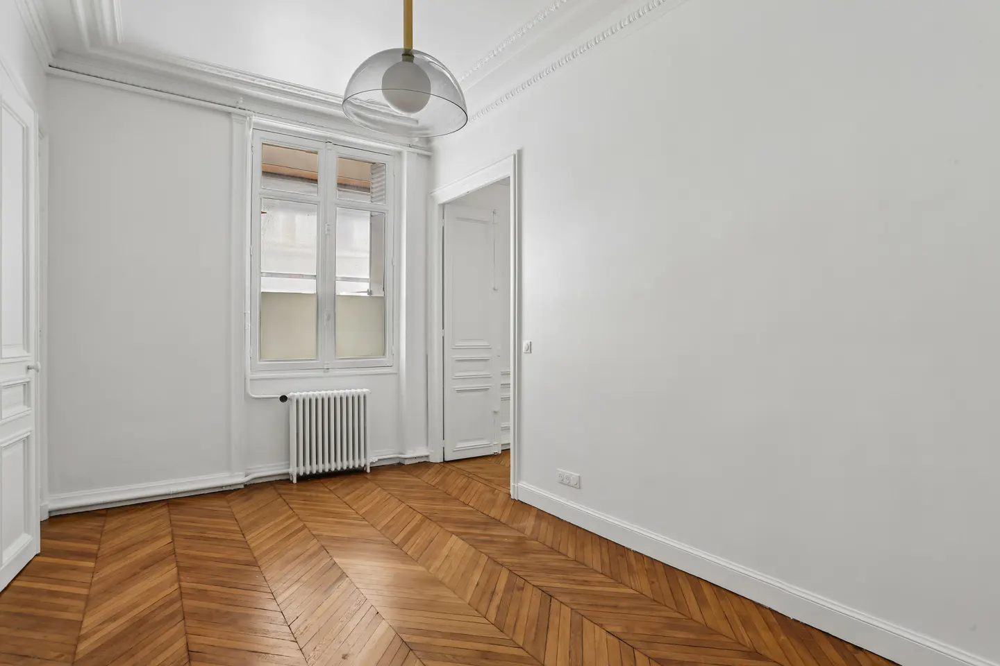 Empty room with white walls, herringbone wood floors, a window with a radiator, and a modern glass pendant light.