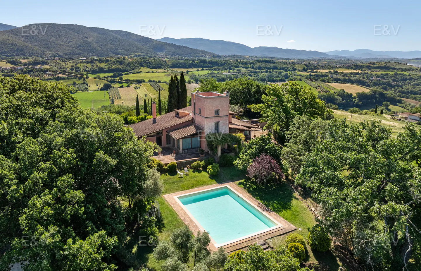 Aerial view of a pink Tuscan villa with a pool, surrounded by lush green trees and rolling hills.