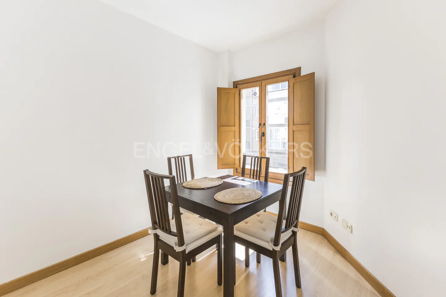 A dining room with a dark wood table and four chairs with white cushions. A window with wooden shutters is in the background.