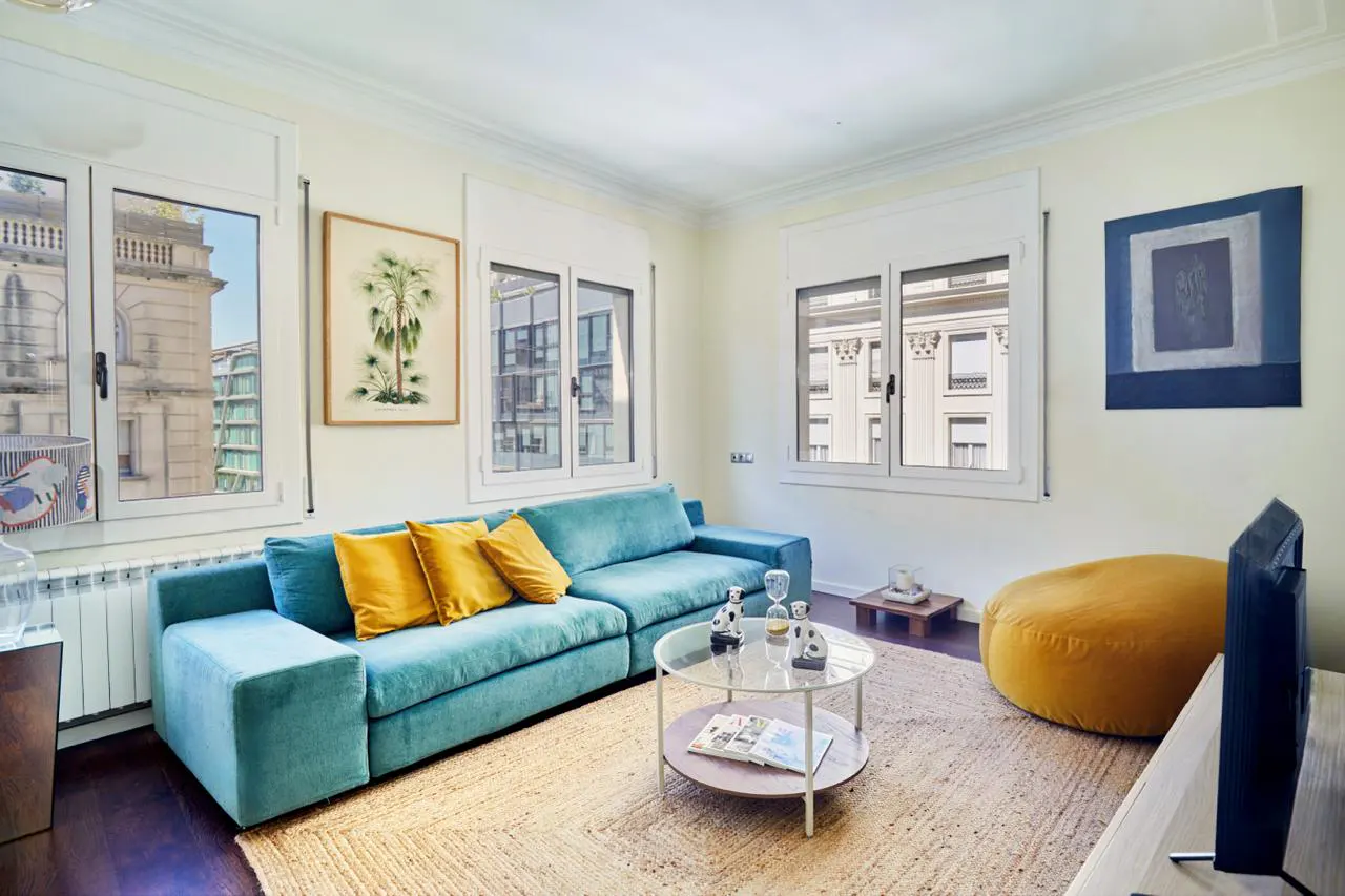 Bright living room with a teal sofa, yellow pillows, and a round glass coffee table on a jute rug. Windows show city views.