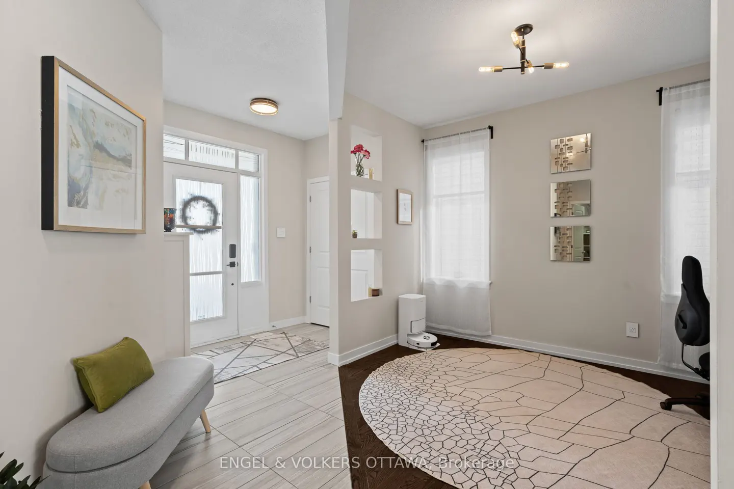 A bright foyer with a white door, a gray bench with a green pillow, and a round rug in the adjacent room.