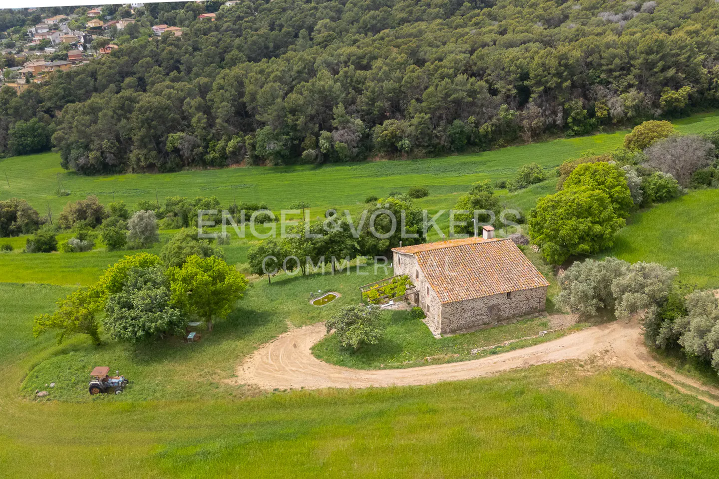 Aerial view of a stone farmhouse with a red tile roof, surrounded by green fields and trees. A dirt road leads to the house.