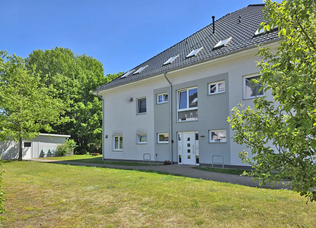 A two-story grey apartment building with a dark tiled roof, skylights, and a white front door, set in a grassy yard.