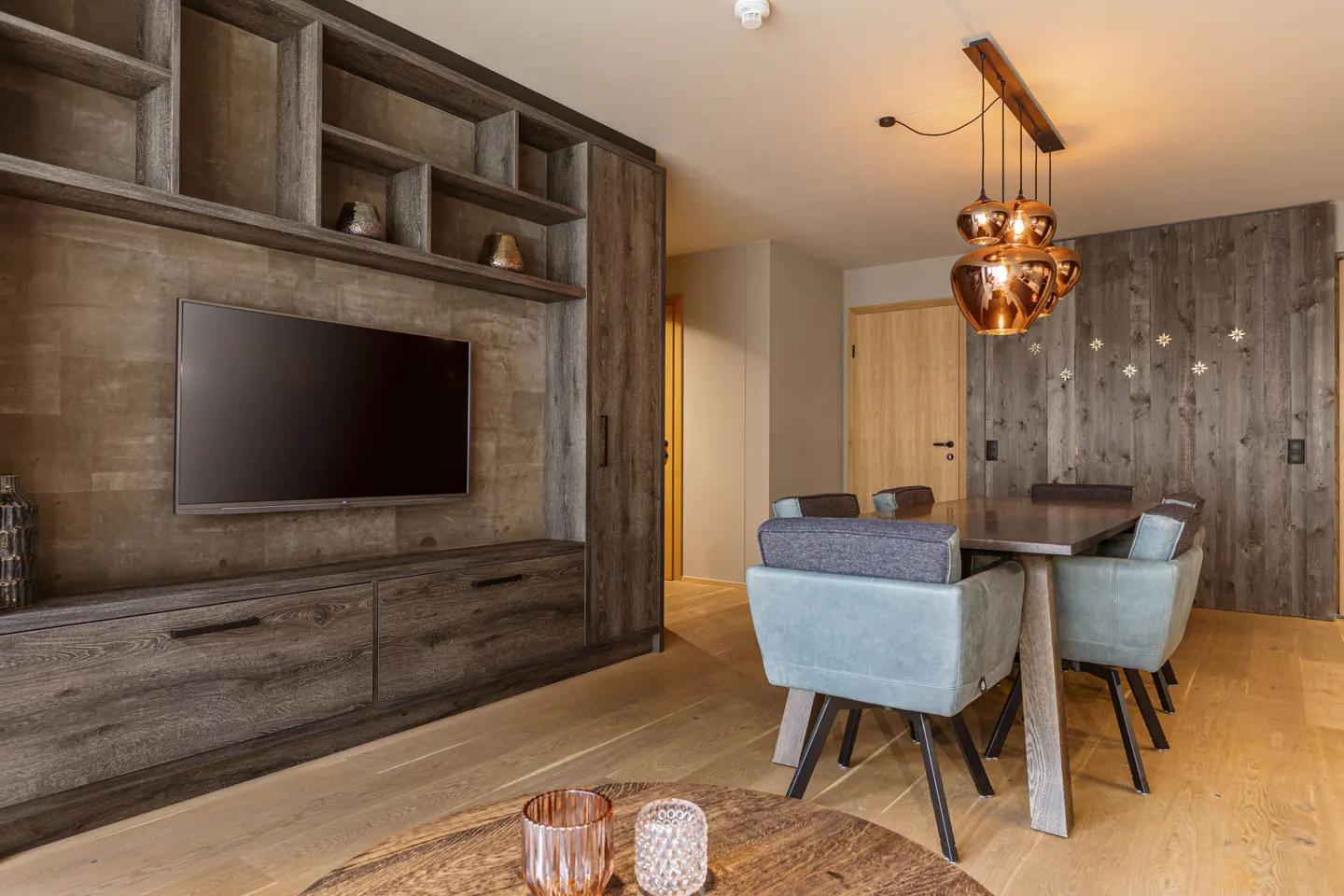 Modern living room with dark wood shelving, wall-mounted TV, dining table with light blue chairs, and copper pendant lights.