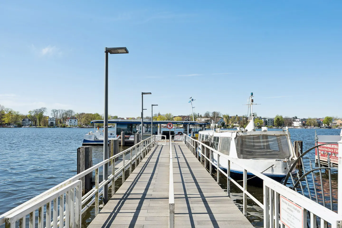A long wooden pier with white railings leads to docked boats on a blue lake under a clear sky.