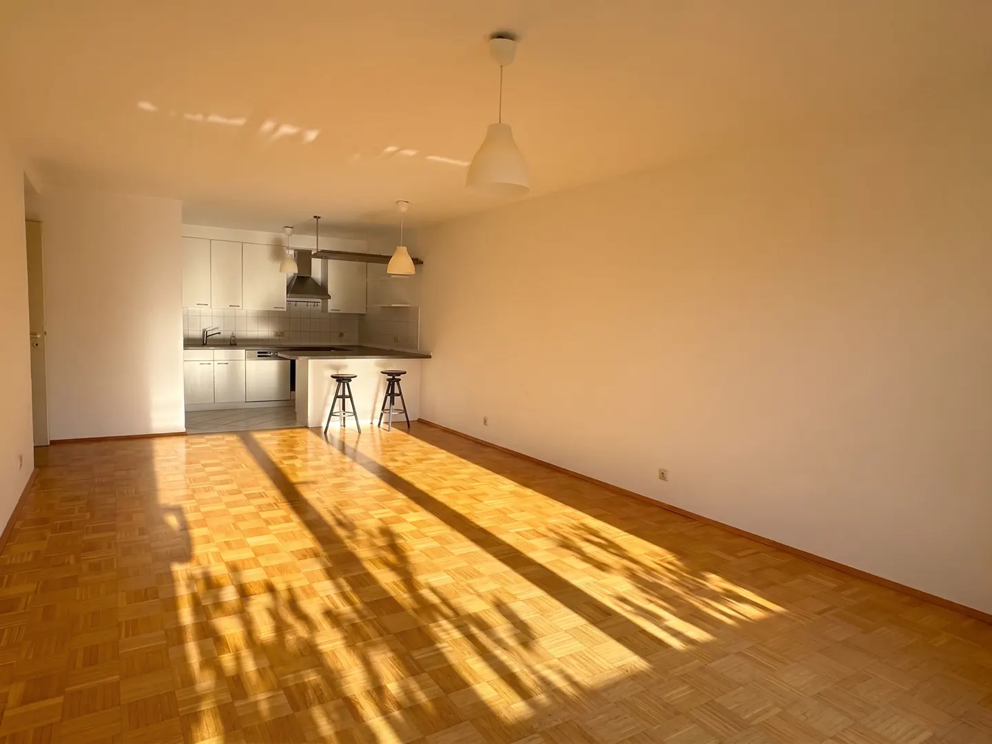 Open-plan living area with light wood parquet flooring, white walls, and a modern kitchen with two bar stools.