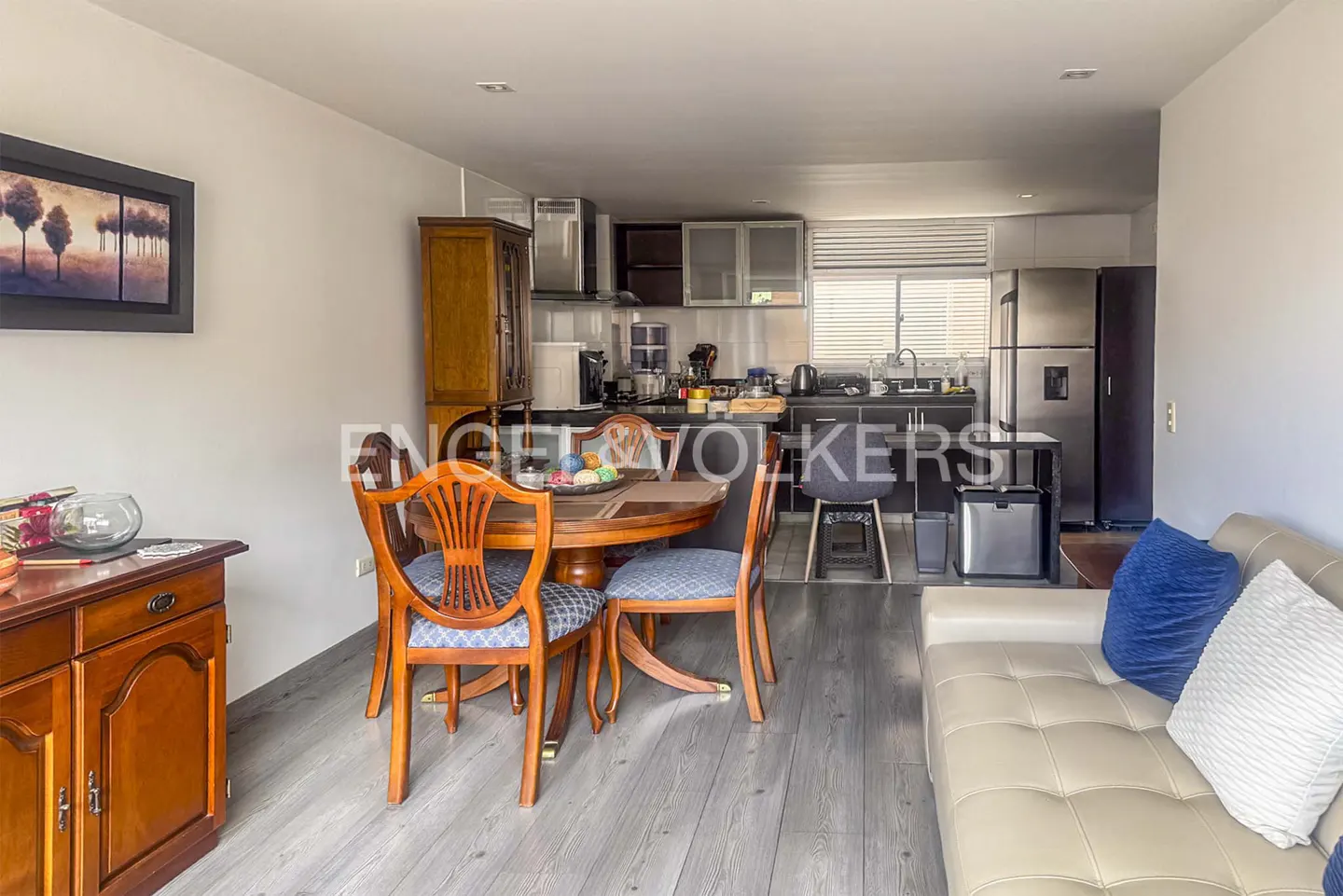 Open-concept living space with a round wooden table, blue-cushioned chairs, and a beige sofa with blue pillows. The kitchen has stainless steel appliances.