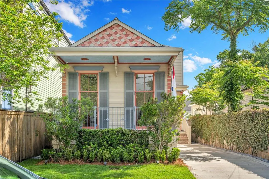 Charming cottage-style home with light yellow siding, pink window trim, and blue shutters, surrounded by lush greenery.