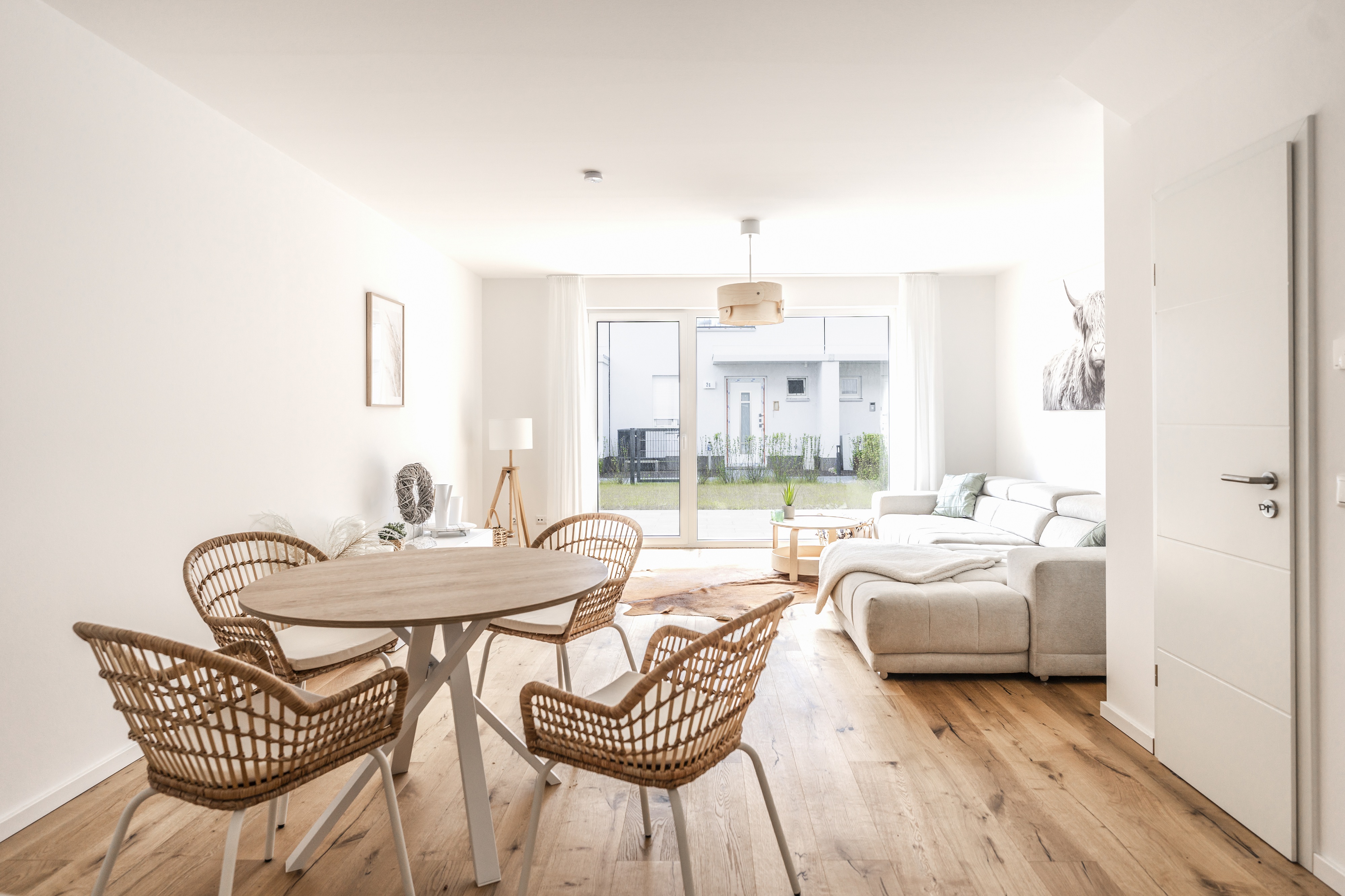 Bright, modern living room with wood floors, a round table with wicker chairs, and a white sofa near a sliding glass door.