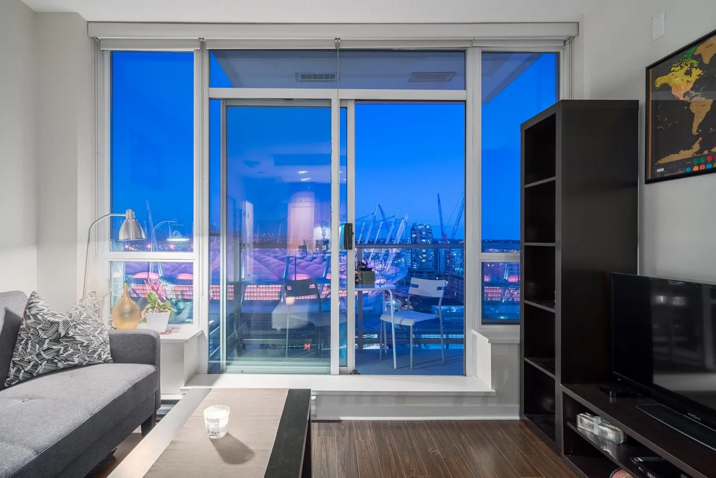 Living room with a gray sofa, dark wood floors, and a balcony overlooking a city skyline at night.