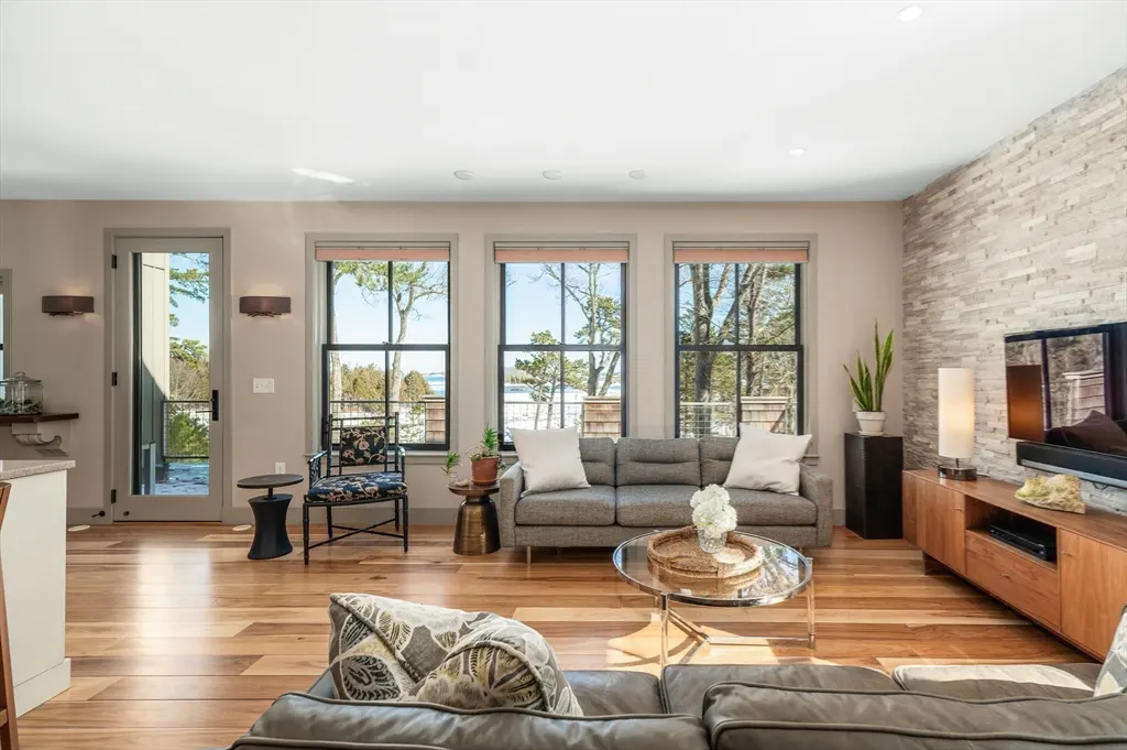 Bright living room with hardwood floors, gray sofa, and large windows overlooking trees and water. A stone accent wall features a flat-screen TV.
