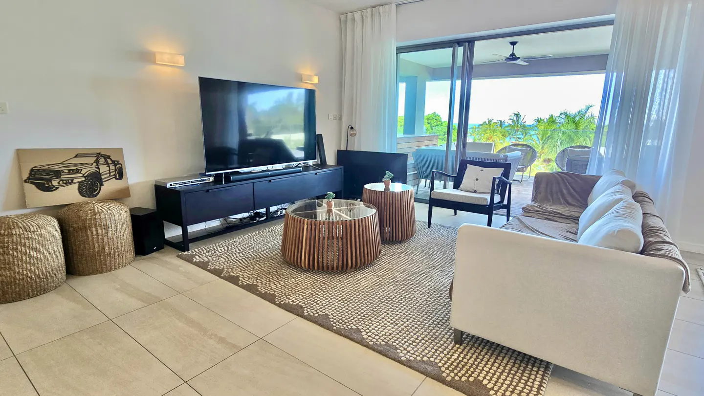 Bright living room with a TV, black console, woven stools, and a white sofa. Sliding glass doors open to a patio with ocean views.