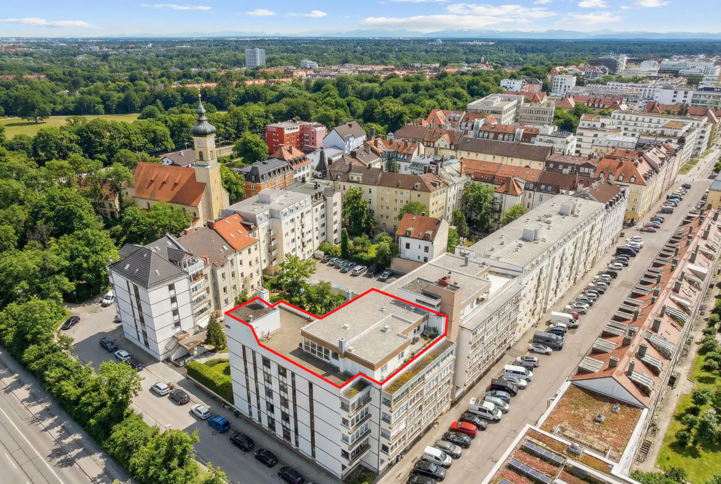 Aerial view of a white apartment building with a red outline, surrounded by trees and other buildings in a city.
