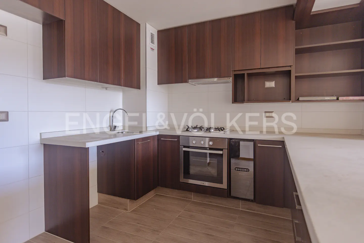 A modern kitchen with dark wood cabinets, white countertops, stainless steel appliances, and light brown tile flooring.