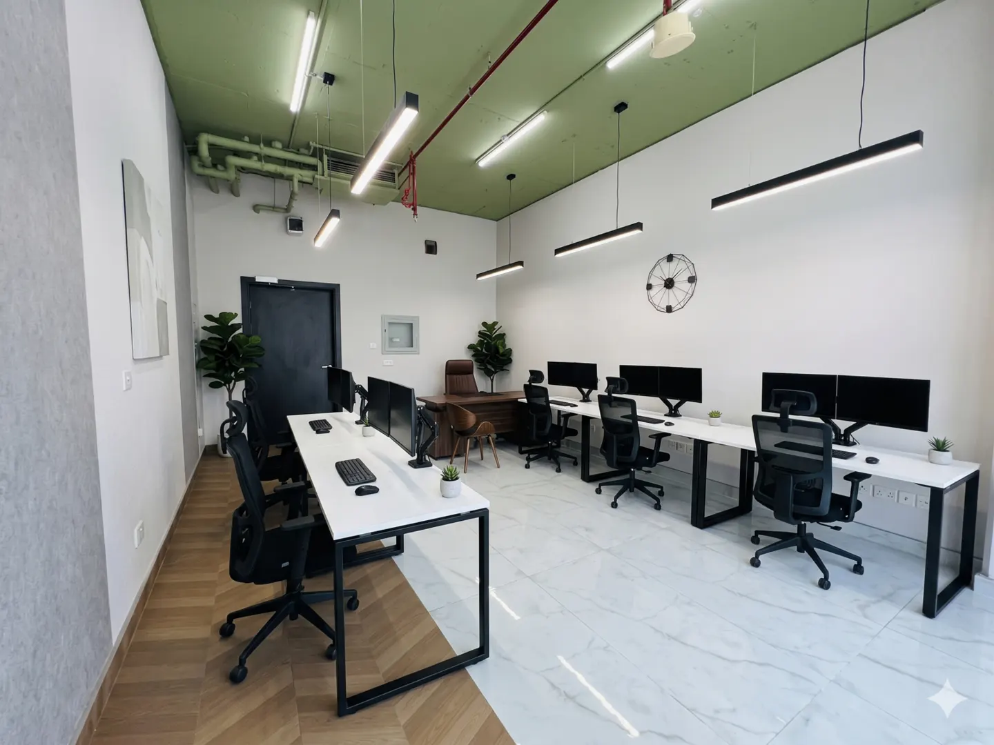 Bright office space with white desks, black chairs, and dual monitors. Green ceiling, linear lighting, and a brown leather executive desk in the background.