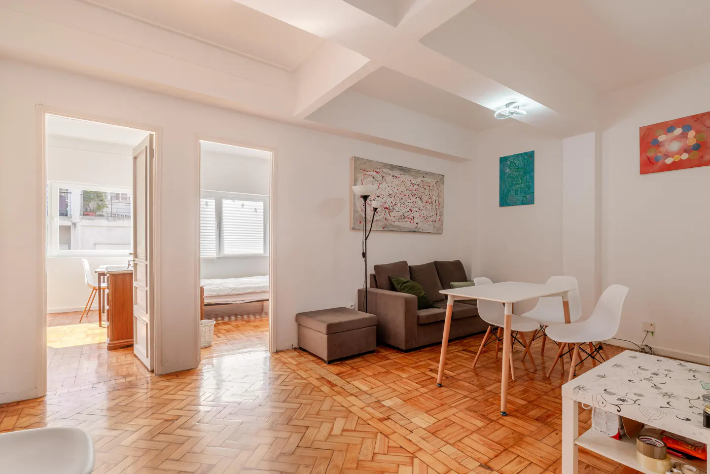 Bright apartment interior with herringbone wood floors, white walls, and modern furniture. A brown sofa and white table sit under abstract art.