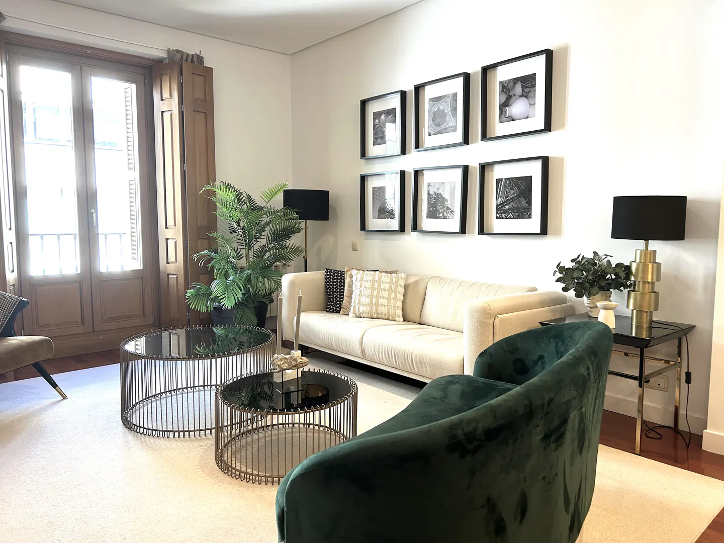 Modern living room with a cream sofa, dark green velvet armchair, two round wire coffee tables, and framed wall art.