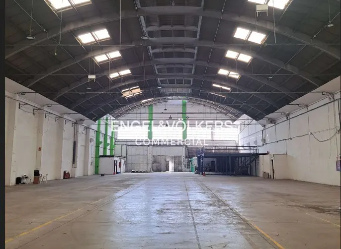 Wide shot of a large, empty warehouse with a curved roof and skylights. The walls are white, and the floor is concrete.