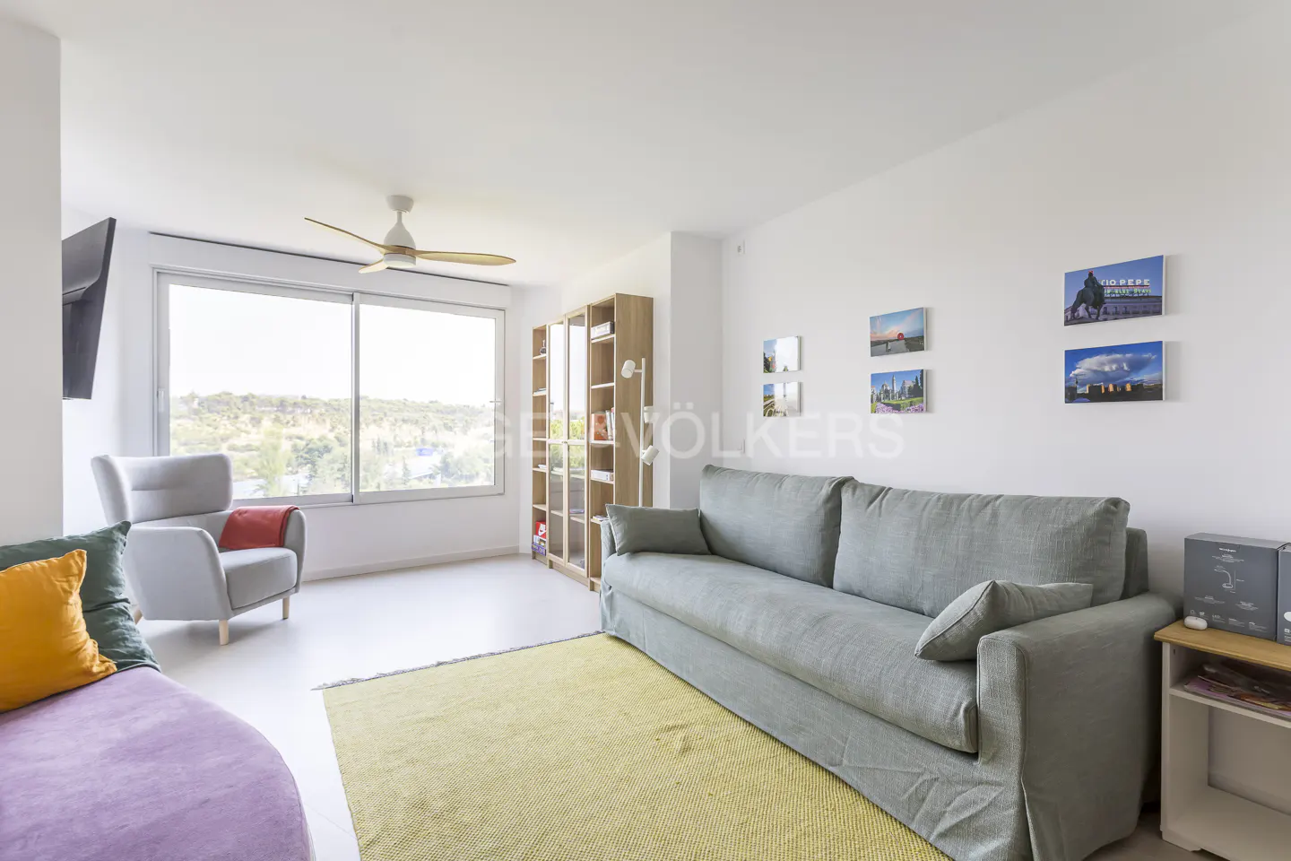 Bright living room with a gray sofa, armchair, and yellow rug. Large window with a view, and photos on the white wall.