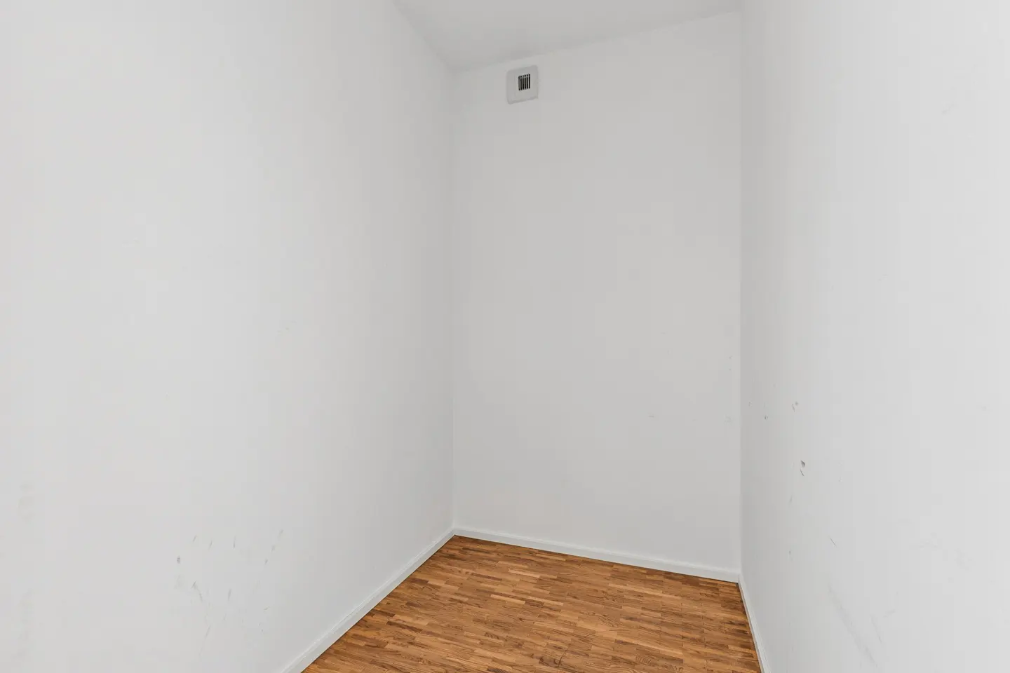 Empty room with white walls and a brown parquet floor. A vent is visible near the ceiling.