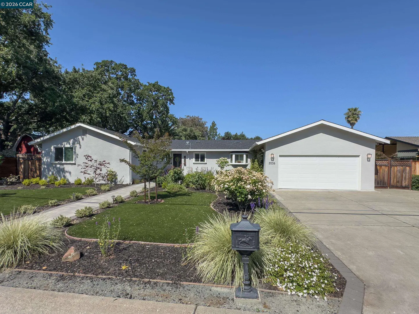 A single-story gray house with a white garage door, green lawn, and black mailbox under a clear blue sky.