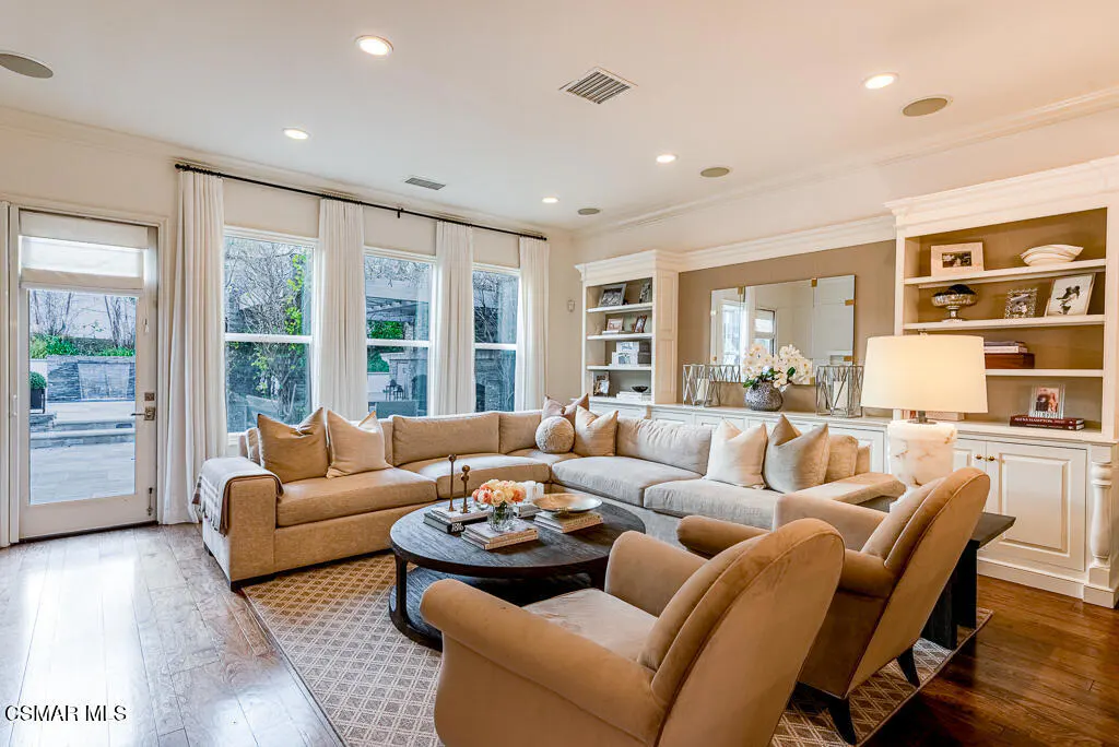 Bright living room with a beige sectional sofa, two armchairs, and a dark wood coffee table on a patterned rug. Built-in shelves and large windows add to the room's appeal.