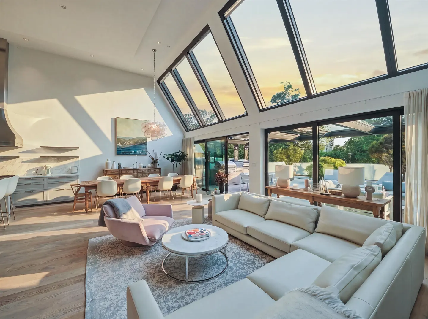 Bright, airy living room with a white sectional sofa, pink chair, and dining area. Large windows offer sunset views.