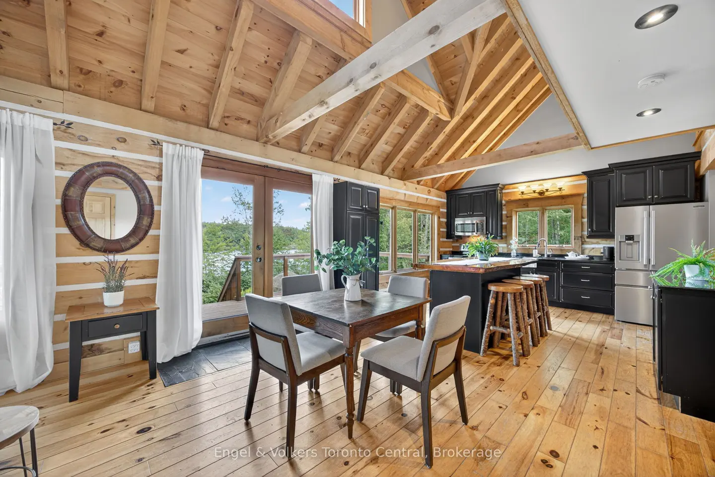Rustic cabin interior with wood walls, floors, and ceiling beams. A dining table sits near patio doors, and a black kitchen is in the background.