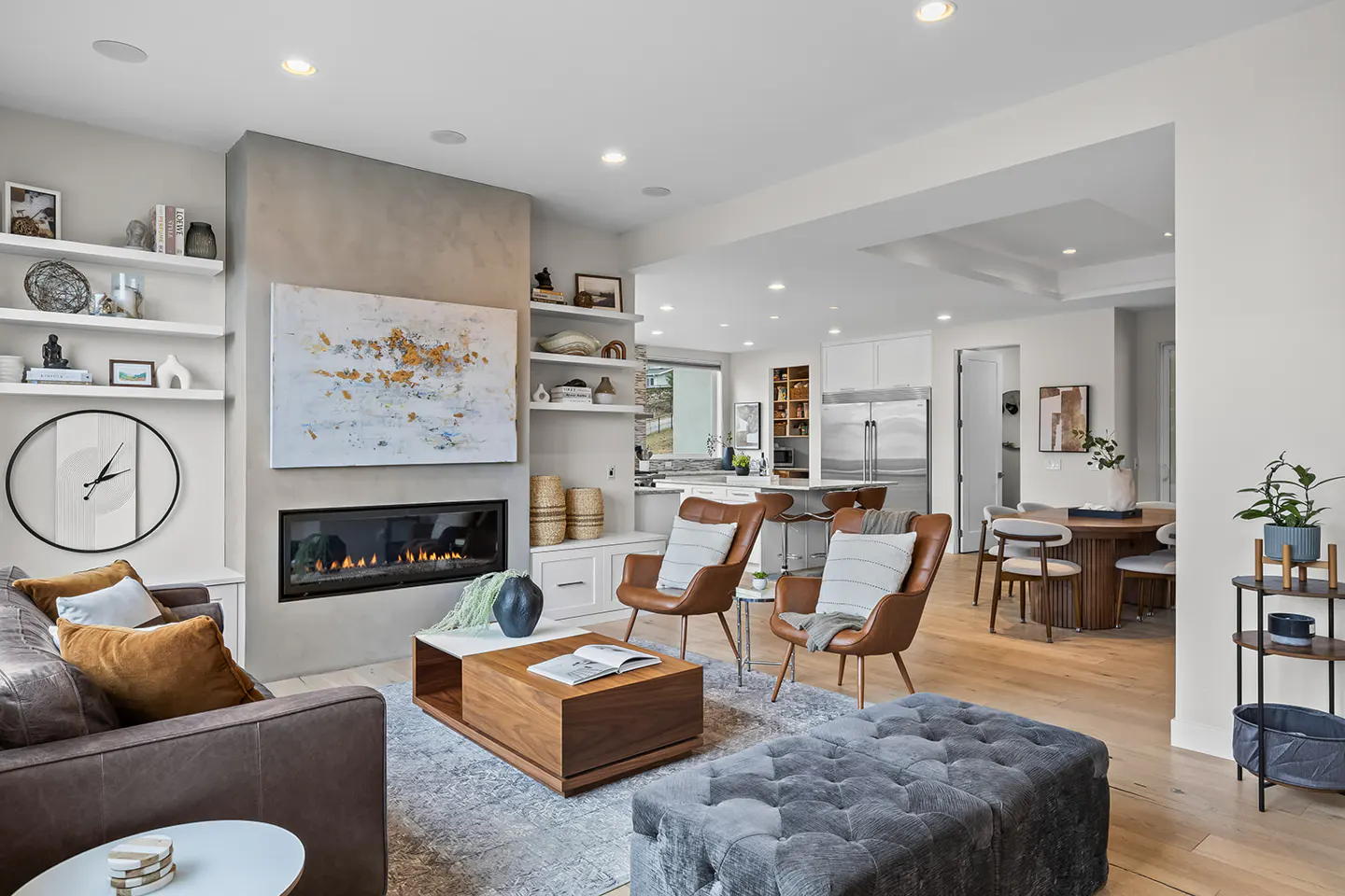 Open-concept living room with a fireplace, brown leather chairs, and a gray tufted ottoman. Kitchen and dining area in the background.
