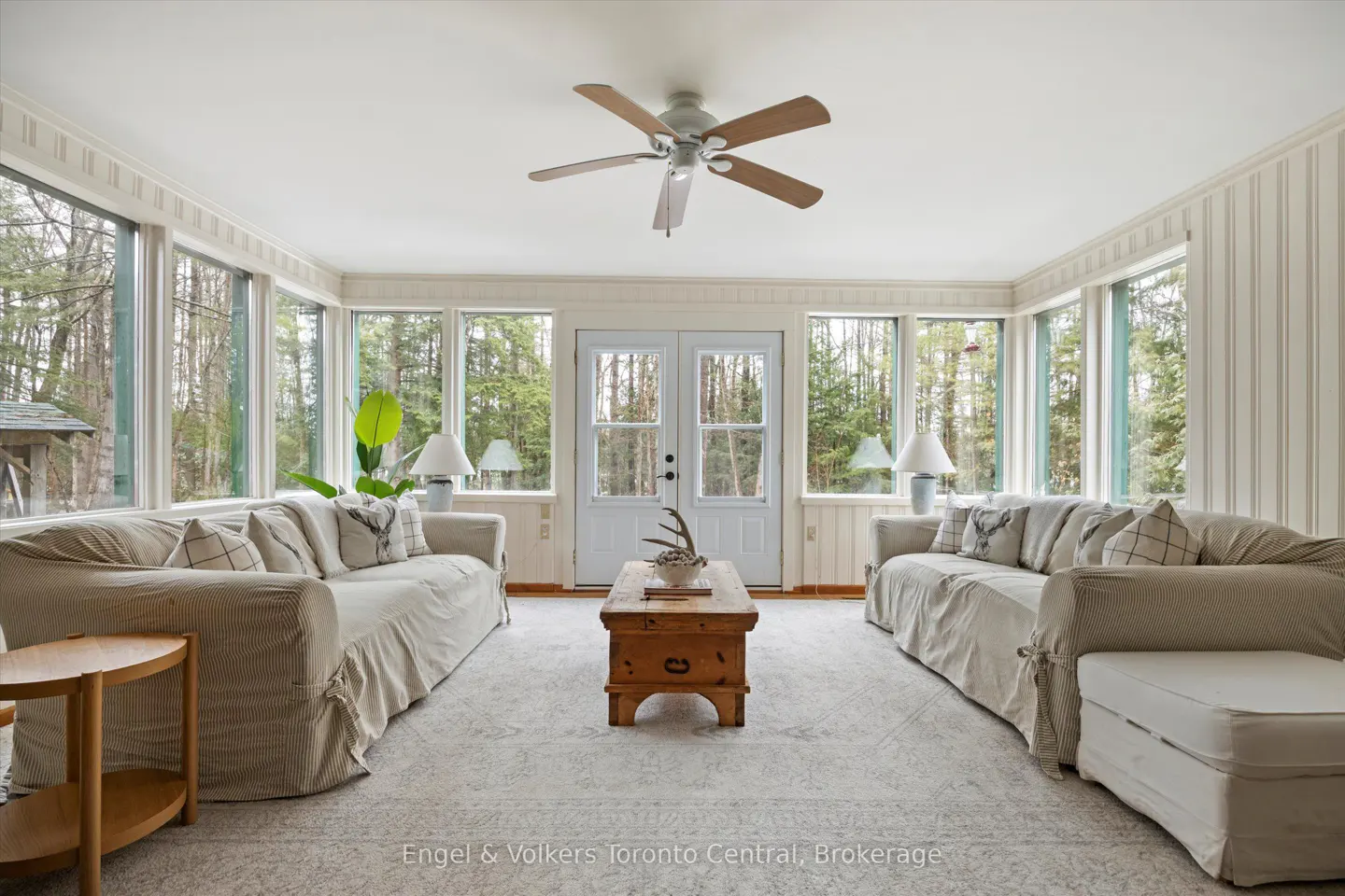 Bright sunroom with white sofas, wood coffee table, and many windows overlooking a forest. A ceiling fan hangs above.