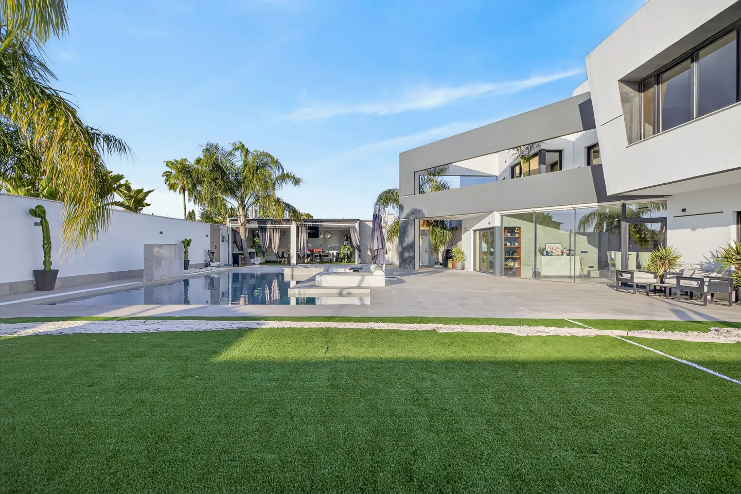 Modern home exterior with a pool, patio, and green lawn under a blue sky. Palm trees and a white wall add to the luxurious outdoor space.