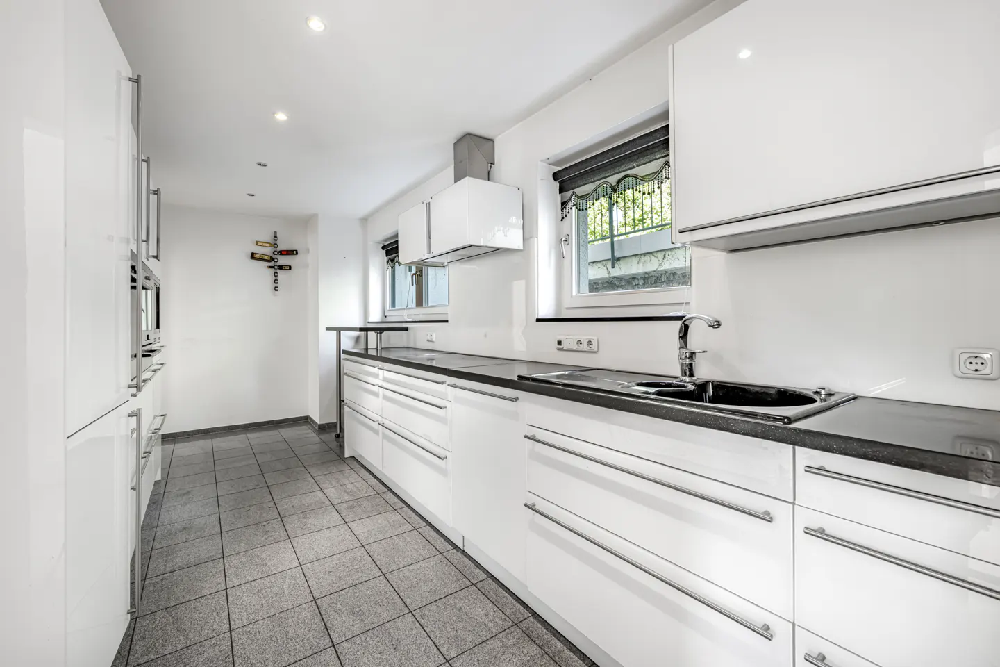 Modern galley kitchen with white glossy cabinets, dark countertops, grey tiled floors, and a wall-mounted wine rack.