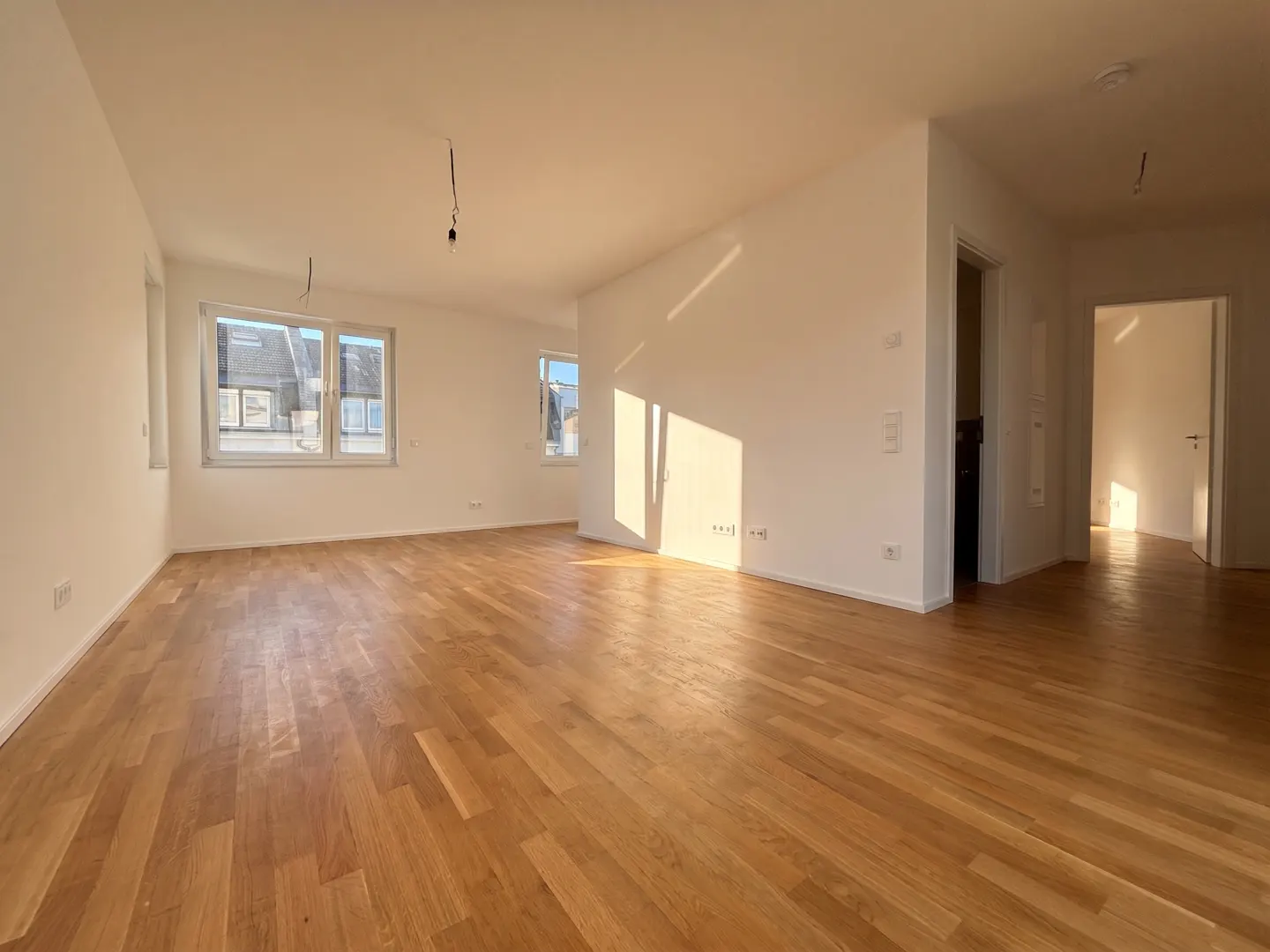 Bright, empty room with light wood floors and white walls. Windows let in sunlight. Doorways lead to other rooms.