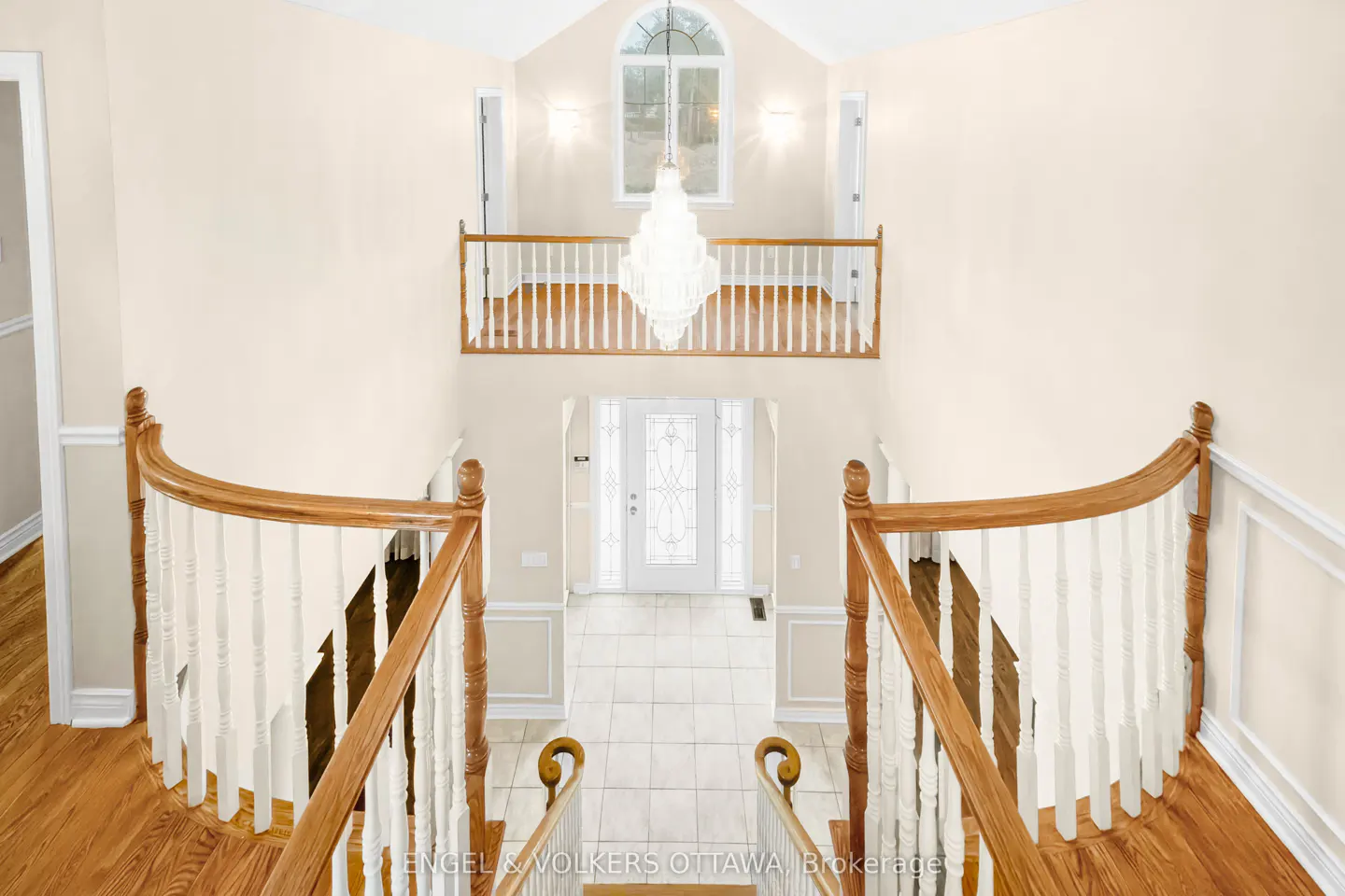 View from the top of wooden stairs looking down to a white front door. A chandelier hangs from the ceiling above a second-floor balcony.