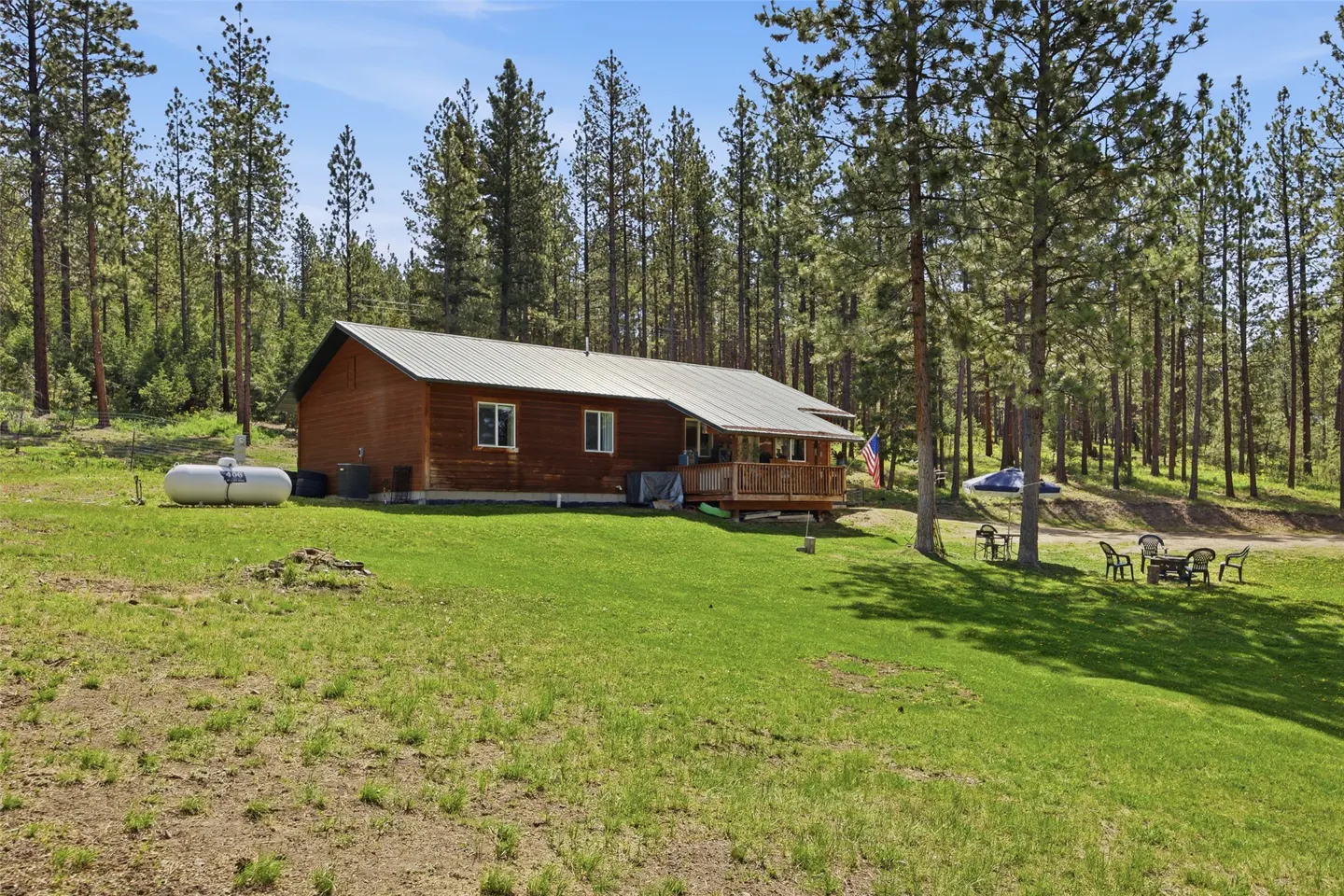 A brown cabin with a metal roof sits on a green lawn surrounded by tall trees. A propane tank is visible on the left.