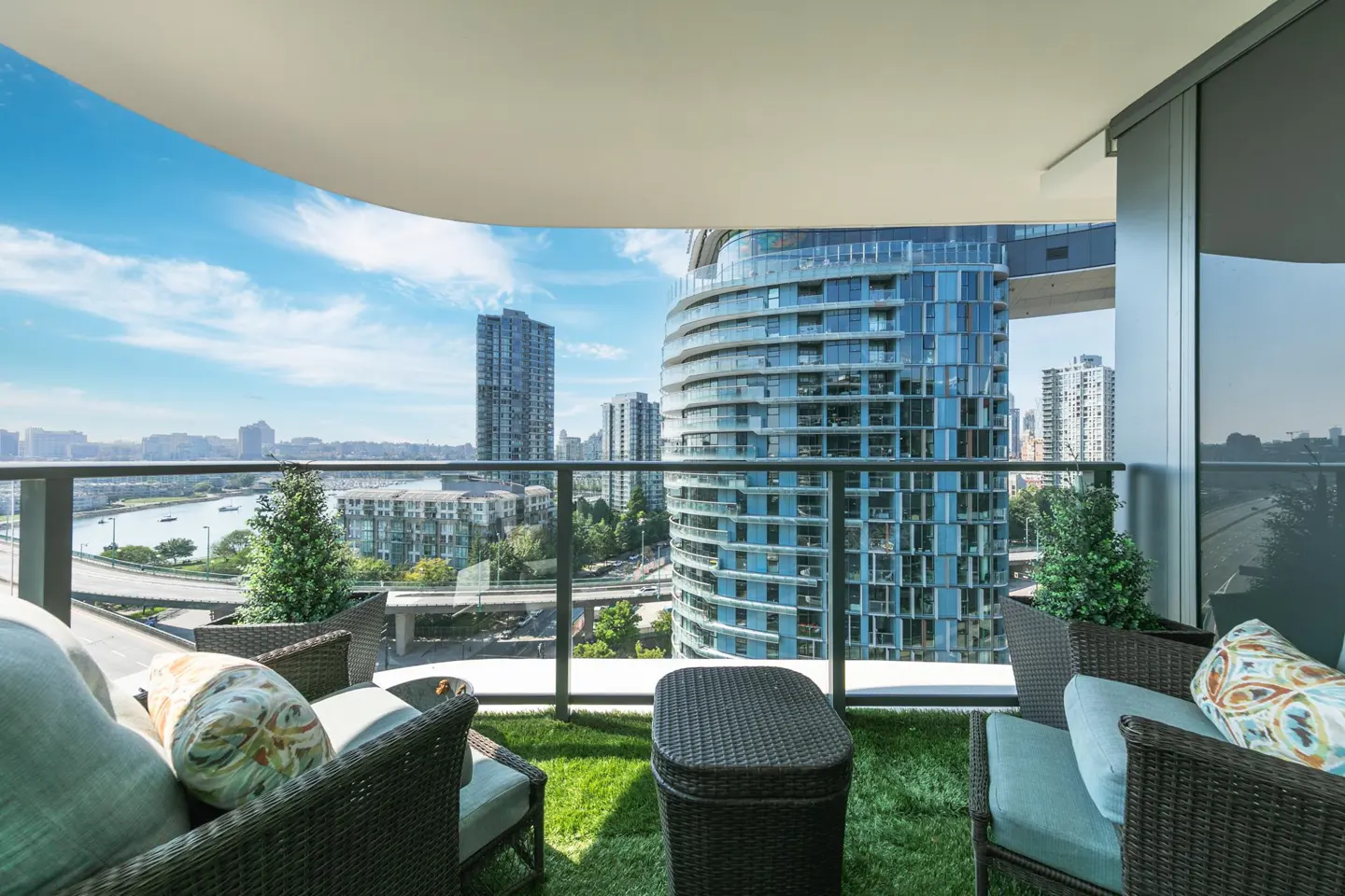 Balcony view with wicker furniture, blue cushions, and city skyline. Tall buildings and a blue sky are visible.