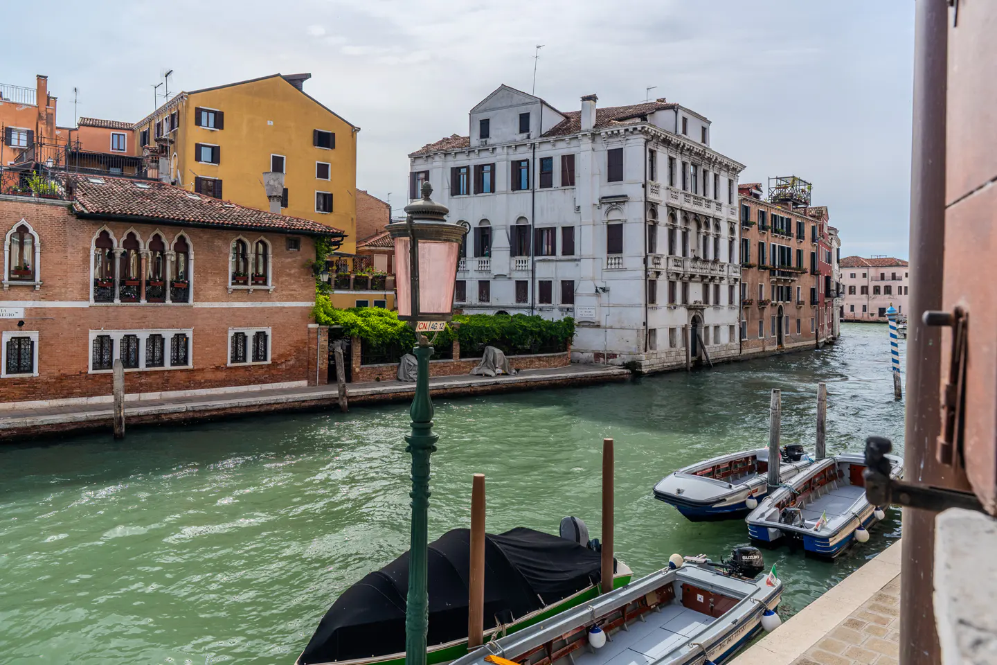 Venice canal view with boats, buildings, and a green lamp post. The water is green, and the sky is cloudy.