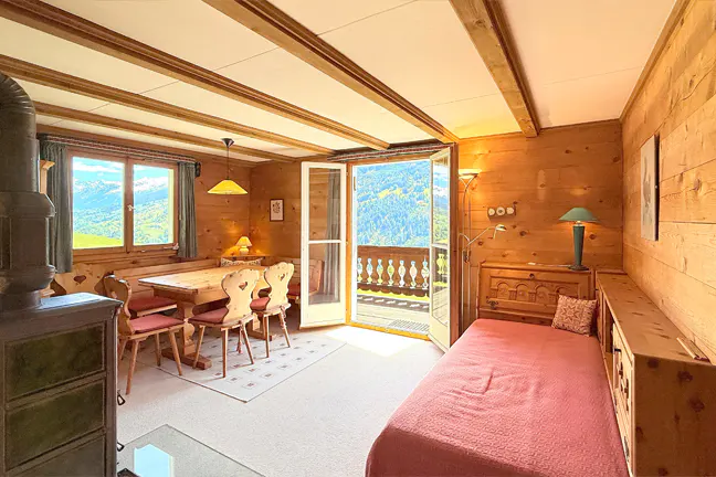 Cozy wood-paneled room with dining table, chairs, and daybed. Balcony doors open to a mountain view. A wood-burning stove is in the corner.