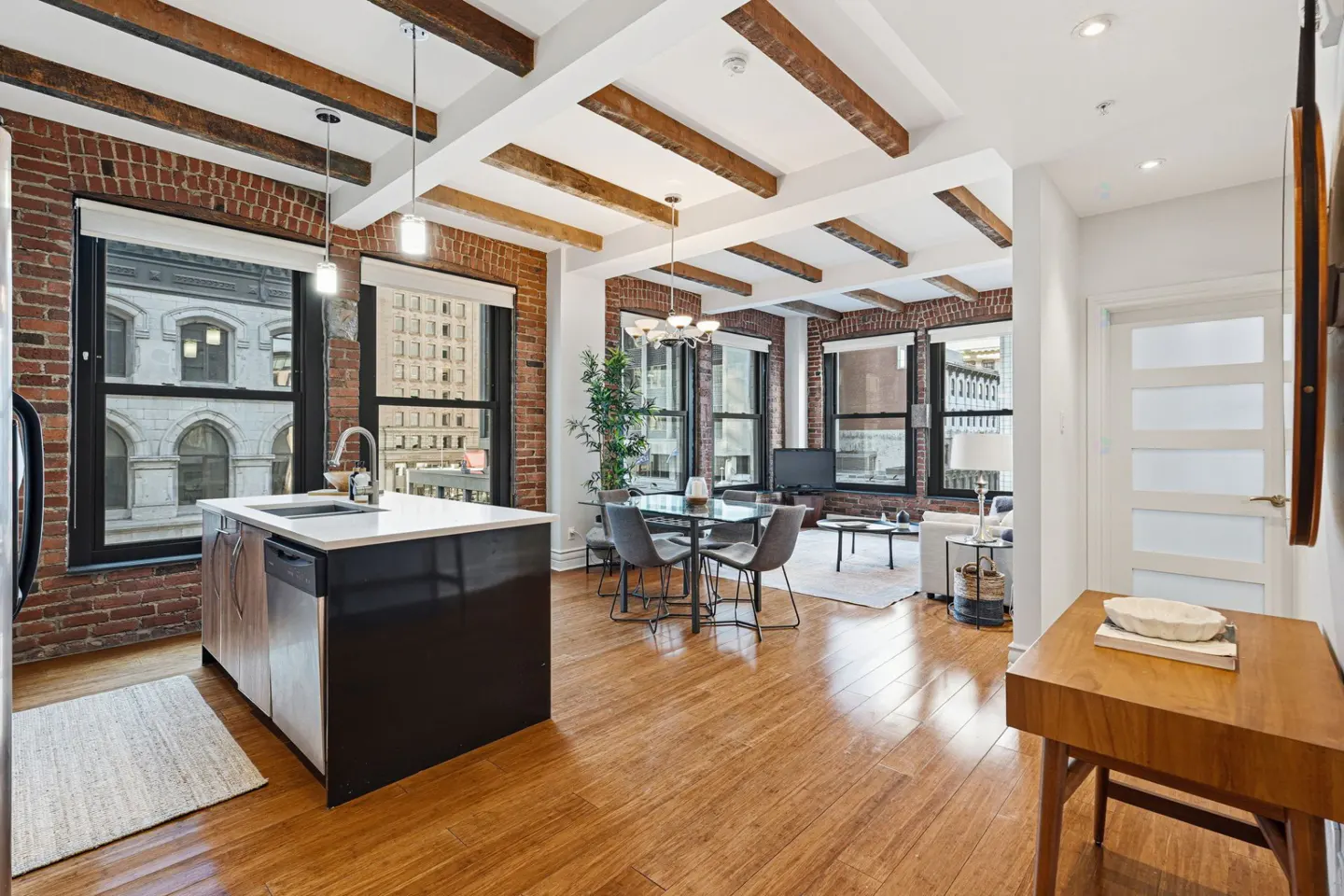 Open-concept loft with exposed brick, wood beams, and hardwood floors. Kitchen island, dining table, and living area visible.