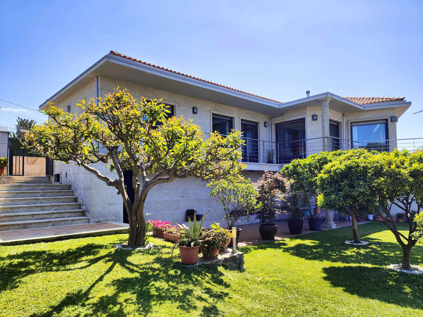 Exterior view of a modern, beige stone house with a red tile roof, green lawn, and trees under a blue sky.